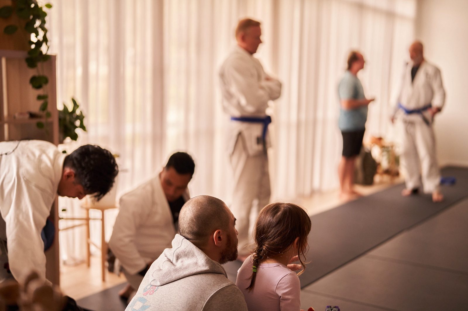 A parent and child sit at the edge of the mats watching training.